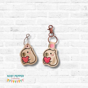 Puede incluir: Dos llaveros beige con un oso de dibujos animados sosteniendo un corazón rosa. Uno tiene una cadena de bolas, el otro un cierre. Los llaveros están bordados con contornos negros y pequeños detalles. El fondo es una pared de ladrillos blancos.