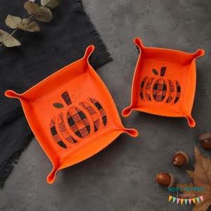 Könnte beinhalten: Zwei leuchtend orangefarbene, quadratische Ablageschalen mit Druckknöpfen an den Ecken, jeweils mit einem gestickten Kürbismotiv in schwarz-orangefarbenem Karomuster. Die Schalen bestehen aus glattem Material und zeigen das Kürbismotiv innen.