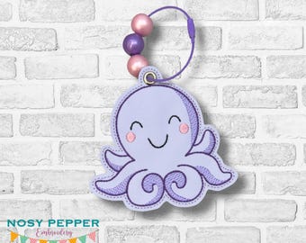 May include: A lavender octopus-shaped keychain with a smiling face and pink cheeks. The keychain has a purple loop and three pink and purple beads. The text "NOSY PEPPER Embroidery" is at the bottom.
