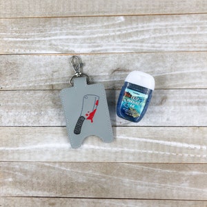 May include: A grey keychain holder with a silver clasp, featuring an embroidered cleaver design with red blood splatter. Beside it is a small bottle of hand sanitiser with the text "CRISP MORNING AIR".