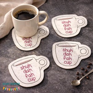 May include: A white ceramic coffee mug filled with dark coffee sits on a cup-shaped coaster. The coaster is beige with a coffee cup design and the words "shuh dah fah cup" in purple. Three additional coasters and a spoon are also visible.