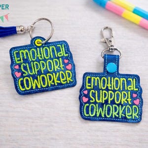 May include: Two blue embroidered keychains with the text "EMOTIONAL SUPPORT COWORKER" in neon green. Each keychain has a silver keyring and a small pink heart design. One keychain has a blue tassel.