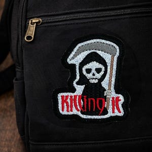 May include: Black backpack with a patch featuring a Grim Reaper with a scythe and the words "Killing It" in red and white. The backpack has a gold zipper and a small pocket.
