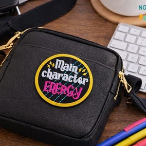 Puede incluir: Bolso bandolera negro con cremallera dorada y un parche redondo que dice "Main character ENERGY" en blanco y rosa. El bolso está sobre una superficie de madera con un teclado, bolígrafos y un bloc de notas.