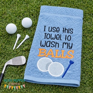 May include: A light blue golf towel with the embroidered text "I use this towel to wash my balls." Two golf balls, two tees, and a golf club are on a green surface.