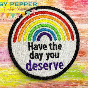 May include: A round white embroidered patch with a rainbow arc and the text "Have the day you deserve" in black and purple lettering. The patch has a black border.