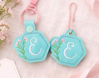 May include: Two turquoise keychains with the letter "E" embroidered in white, surrounded by pink floral accents. Each keychain has a pink clasp or loop. The keychains are hexagonal and have a stitched border.
