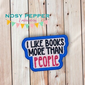 Puede incluir: Parche bordado azul y blanco con el texto "Me gustan más los libros que las personas" en letras negras y rosas.