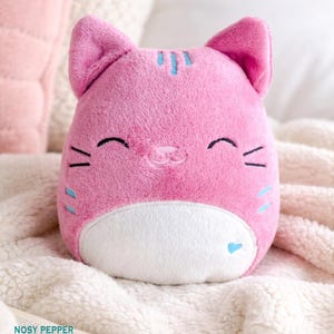 Könnte beinhalten: Ein rosa Plüsch-Katzenspielzeug mit weißem Bauch, blauen Akzenten und gestickten Gesichtszügen. Die Katze hat ein lächelndes Gesicht und liegt auf einer weichen, weißen Decke. Das Spielzeug ist ein niedliches, kuscheliges Objekt.