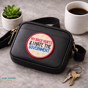 May include: Black crossbody bag with a patch that reads "MY BACK HURTS & I HATE THE GOVERNMENT". The bag has a gold chain and a black strap. A small succulent in a white pot and a cup of coffee are in the background.