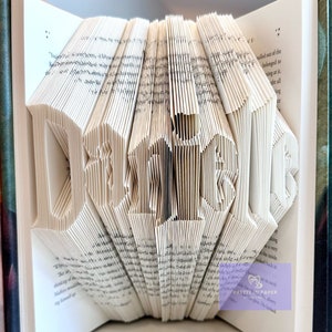 Peut inclure: Une sculpture d'art de livre du mot "Dumbledore" créée en pliant les pages d'un livre. Le livre est ouvert et éventailé pour former les lettres. Le livre est entouré de deux autres livres, dont un avec le titre "Harry Potter et la Coupe de Feu" visible.
