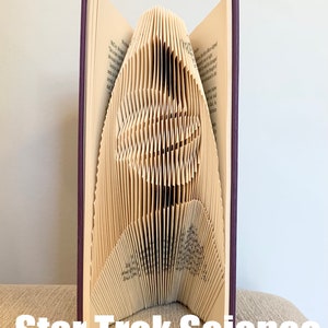 May include: A book with pages folded into a three-dimensional sculpture of the Star Trek insignia. The book is closed and has a purple cover. The sculpture is standing on a beige surface. The text "Star Trek Science" is printed below the sculpture.