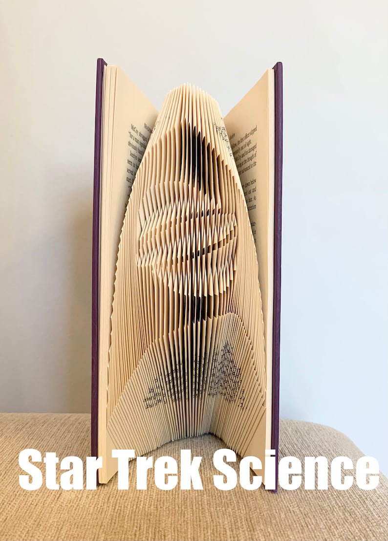May include: A book with pages folded into a three-dimensional sculpture of the Star Trek insignia. The book is closed and has a purple cover. The sculpture is standing on a beige surface. The text "Star Trek Science" is printed below the sculpture.