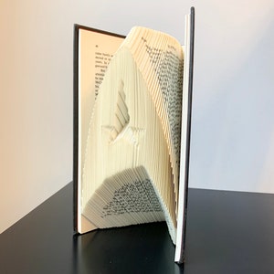 May include: A book art sculpture of a bird made by folding the pages of a book. The book is open and the pages are folded into a three-dimensional shape. The sculpture is standing on a black surface.