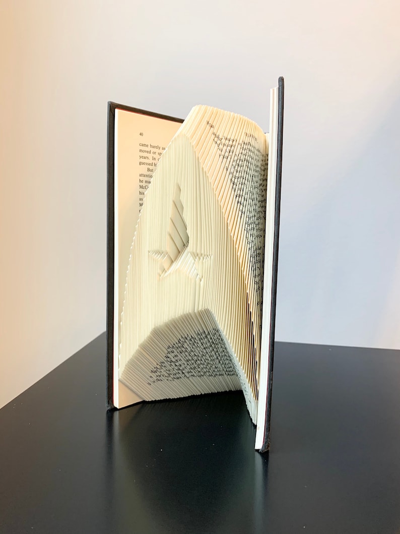 May include: A book art sculpture of a bird made by folding the pages of a book. The book is open and the pages are folded into a three-dimensional shape. The sculpture is standing on a black surface.
