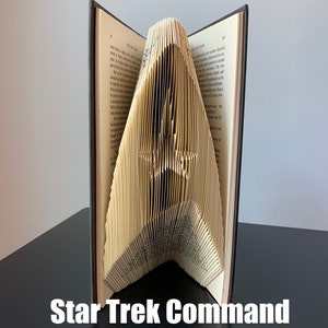 May include: A book folded into the shape of the Star Trek command insignia. The book is open and the pages are folded to create the iconic shape. The book is sitting on a dark surface. The text "Star Trek Command" is written below the book.
