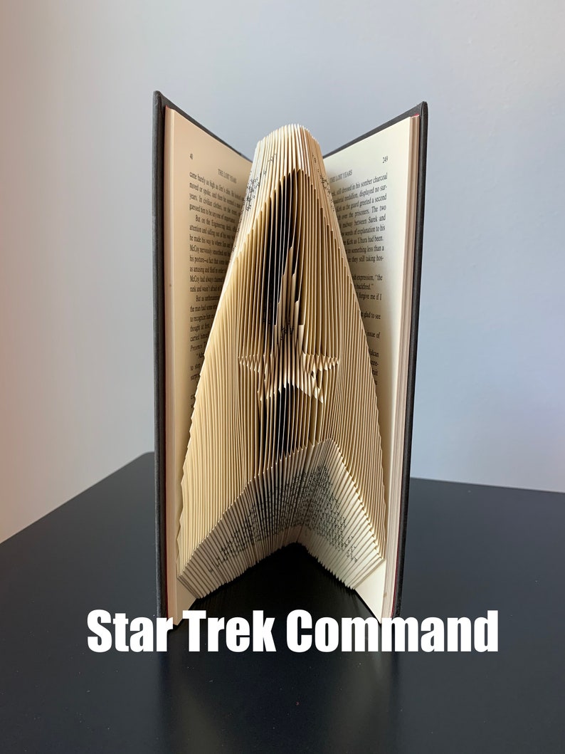 May include: A book folded into the shape of the Star Trek command insignia. The book is open and the pages are folded to create the iconic shape. The book is sitting on a dark surface. The text "Star Trek Command" is written below the book.