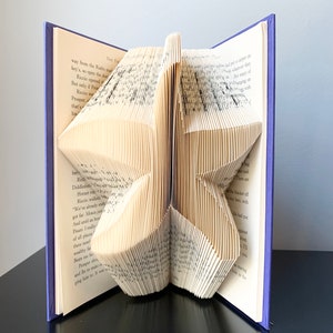 May include: A book with pages folded into a 3D star shape. The book is bound in a purple cover and is sitting on a black surface.