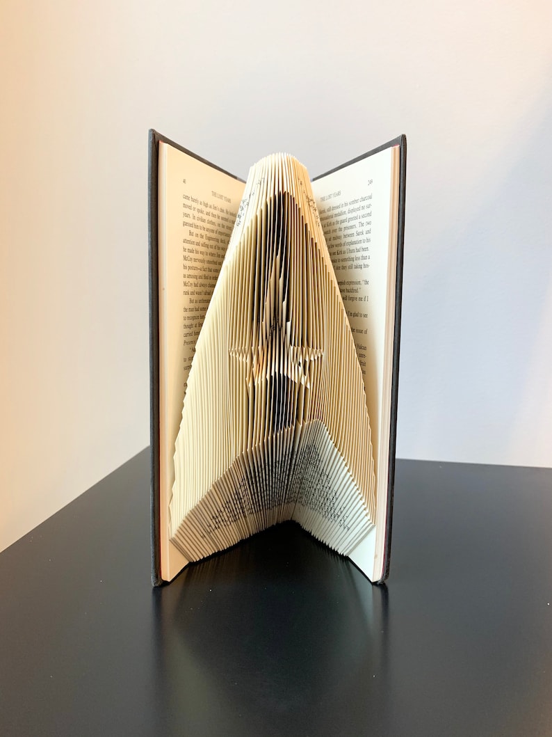 May include: A book with pages folded into a three-dimensional shape, creating a sculpture of the letter "A". The book is open and the sculpture is standing on a black surface.