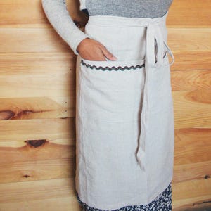 May include: A white linen apron with a decorative trim of blue and green embroidery. The apron has a large pocket on the front and ties at the waist.