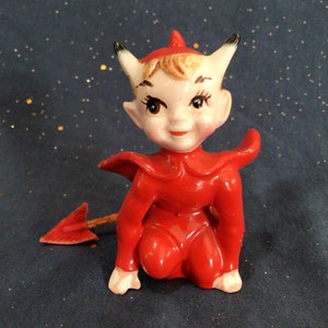 May include: A small ceramic figurine of a red devil with a pointed tail. The devil is wearing a red suit and has a mischievous expression on its face.