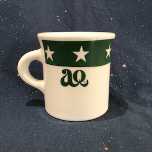 May include: White ceramic mug with a green band and white stars. The mug has the letters "aQ" in green on the front.