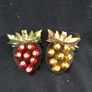 May include: Two Christmas ornaments shaped like strawberries with gold foil leaves. One ornament has red berries with white centers and the other has gold berries with white centers.