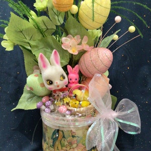 May include: A vintage-style Easter centerpiece with a pink and white bunny, yellow chicks, and pastel-colored eggs in a decorated tin can with a pink bow.