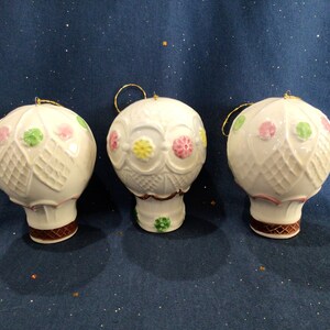 May include: Three white ceramic hot air balloon ornaments with colorful floral designs. The ornaments have a gold string for hanging.