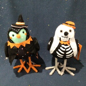 May include: Two Halloween-themed felt bird ornaments. One is a black bird wearing a witch's hat with a green face and orange legs. The other is a white bird with a skeleton costume and orange legs.