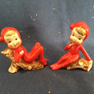 May include: Two ceramic figurines of elves in red hooded jumpsuits, resting on brown tree stumps. The elves have fair skin, blonde hair, and are in relaxed poses. The background is a dark blue with gold flecks.