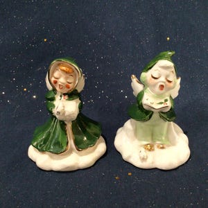 May include: Two vintage porcelain Christmas angel figurines. The angels are dressed in green robes with gold trim and have white wings. They are standing on white clouds.