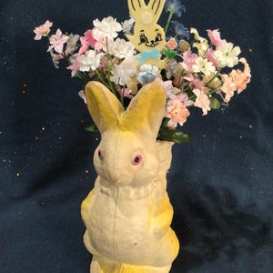 May include: A vintage Easter bunny figurine, painted yellow and white, holding a bouquet of pastel-colored artificial flowers. The bunny has pink eyes and a small yellow bunny on top. A charming, nostalgic Easter decoration.