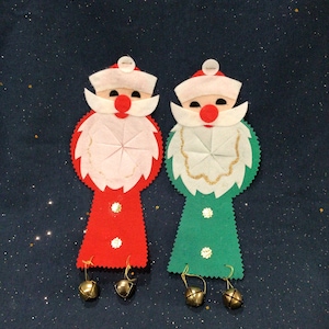 May include: Two felt Christmas ornaments shaped like Santa Claus. The Santa on the left is wearing a red suit and the Santa on the right is wearing a green suit. Both Santas have white beards and white hats with red pom-poms. Each Santa has a gold jingle bell hanging from their feet.