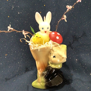 May include: A ceramic Easter bunny planter with a yellow chick and a red mushroom. The planter is decorated with green leaves and a brown base.