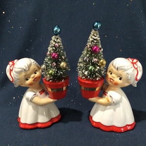 May include: Two ceramic figurines of girls in white dresses with red trim, each holding a small Christmas tree in a red pot. The trees are decorated with colorful ornaments and topped with a blue ball. The background is a dark blue.