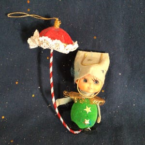 May include: A vintage Christmas ornament featuring a doll-like figure with a green body adorned with gold stars. The doll wears a white hat and holds a red and white striped umbrella with a red top and white lace trim. The ornament has a gold string for hanging.
