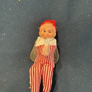 May include: A small doll with a peach-colored face, blue eyes, and a red cap, wearing a red and white vertically striped outfit with a white collar. The doll has translucent gold-toned wings and is holding a small white candle in its hands.