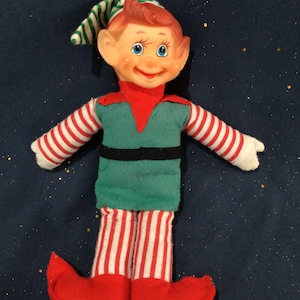 May include: A vintage elf doll with a painted face, red hair, and blue eyes. The elf is wearing a green and red outfit with striped sleeves and legs, a black belt, and red shoes. The elf has a green and white striped hat.