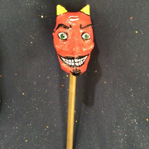 May include: A red devil-faced lollipop with yellow horns, green eyes, and a black mustache and goatee. The lollipop is on a wooden stick. The background is a dark blue surface with gold glitter.