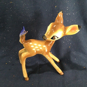 May include: A ceramic figurine of a fawn with brown and white spots. The fawn is standing on all four legs and has its head turned to the side. The figurine is painted with a blue and brown color scheme.