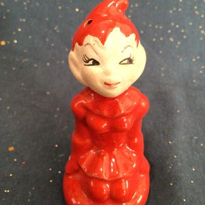May include: A red ceramic elf figurine with a white face and black eyes. The elf is sitting with its legs crossed and has a pointed hat.