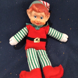 May include: A vintage elf doll with a red and green outfit. The doll has a red tunic, red shoes, and green and white striped legs and sleeves. The elf has a green hat with red and white stripes and a black belt with a gold buckle.