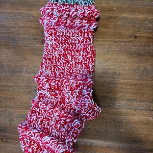 Puede incluir: Un calcetín de Navidad de crochet rojo y blanco con un puño de crochet verde. El calcetín tiene un borde rizado y está decorado con pompones blancos.