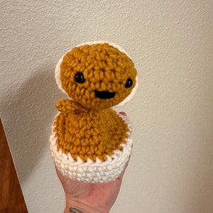 Puede incluir: Un juguete de peluche tejido a crochet con forma de persona, con un cuerpo marr&oacute;n y un gorro blanco tejido a crochet. El juguete tiene ojos negros y una boca sonriente.