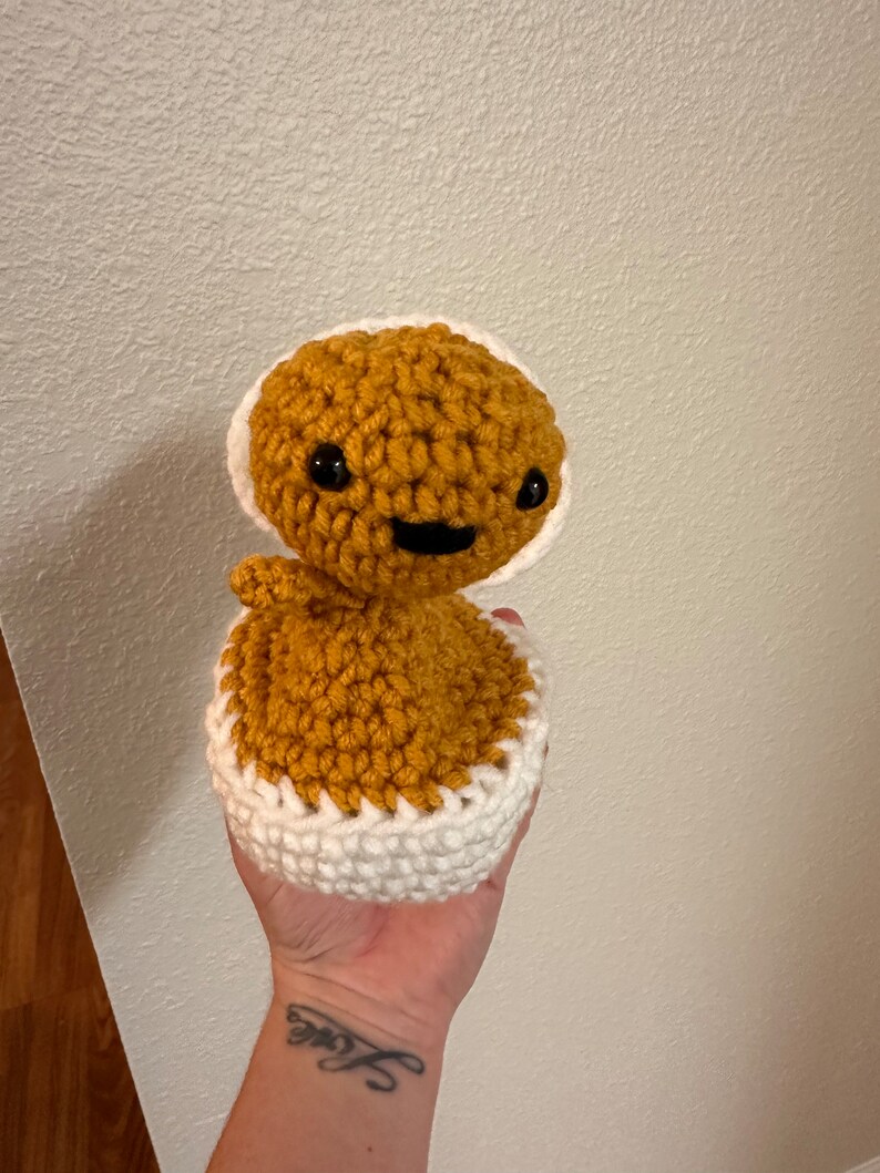 Puede incluir: Un juguete de peluche tejido a crochet con forma de persona, con un cuerpo marr&oacute;n y un gorro blanco tejido a crochet. El juguete tiene ojos negros y una boca sonriente.