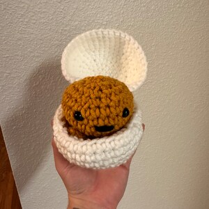 Puede incluir: Un juguete amigurumi de crochet con forma de criatura marr&oacute;n con una cara sonriente, dentro de una concha de crochet blanca.