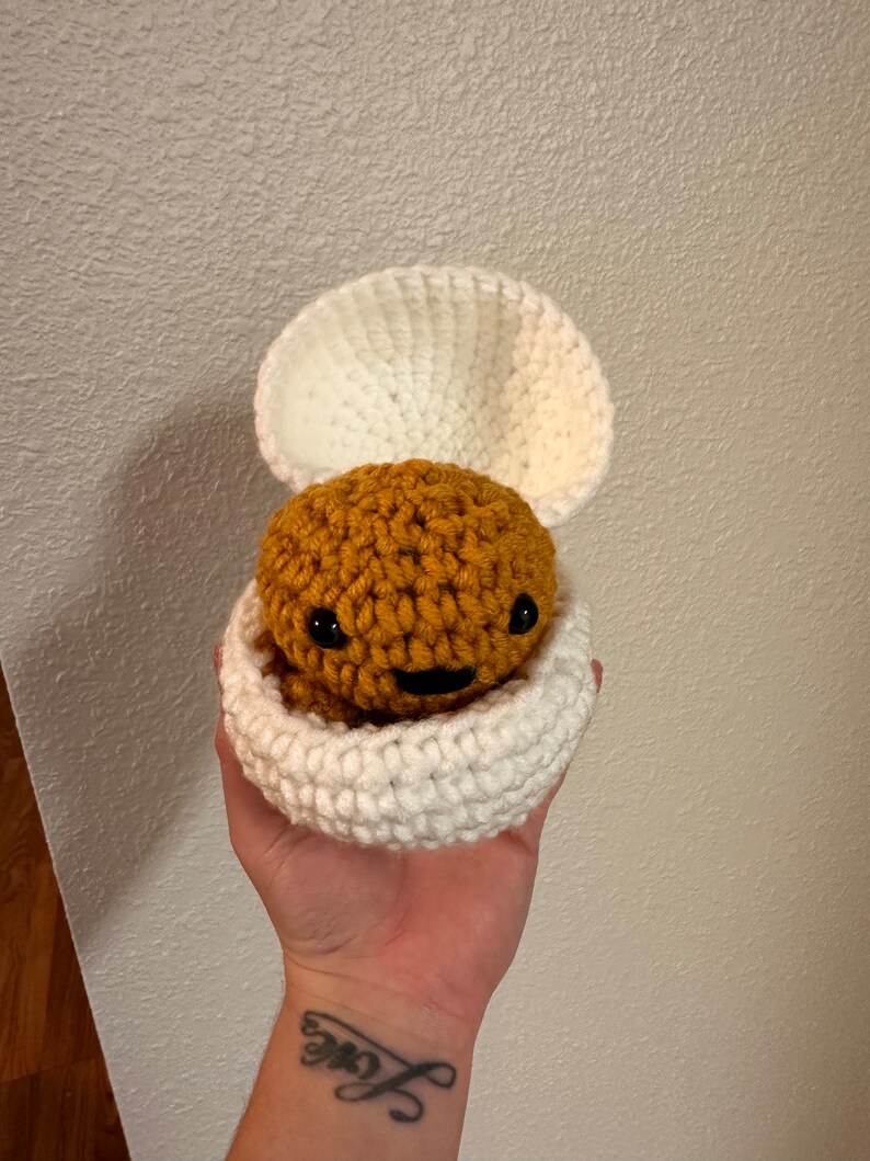Puede incluir: Un juguete amigurumi de crochet con forma de criatura marr&oacute;n con una cara sonriente, dentro de una concha de crochet blanca.