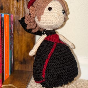 Puede incluir: Una muñeca de ganchillo hecha a mano con piel beige claro, pelo de lana marrón y un sombrero rojo oscuro. La muñeca lleva un vestido negro con un borde rojo en el centro y una capa negra. Tiene dos pequeños ojos de cuentas negras.