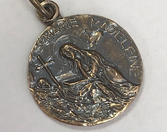 Könnte beinhalten: Bronzefarbener Anhänger mit der Inschrift "St. Marie Madeleine". Der Anhänger zeigt ein detailliertes Relief einer knienden Figur mit Kreuz und Heiligenschein. Er hat eine kleine Schlaufe oben zum Befestigen an einer Kette.